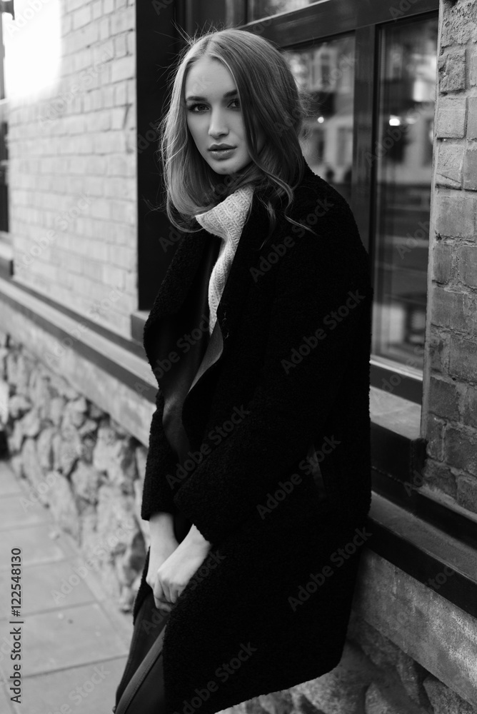 Black and white photo. Beautiful young blonde woman walking on the street. Dressed in a black coat and a warm knit sweater.