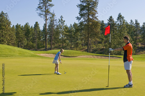 A junior female golfer gets ready to putt while her father readies the flagstick on a green at Two Eagles Golf Course in Westbank, British Columbia, Canada photo