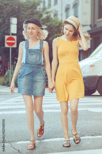 Portrait of two white Caucasian unformal young girls hipster students teenagers friends in dresses  hats  outside running crossing urban city street crowded with cars  best friends forever