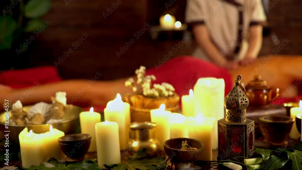 Still life with burning candles and massage herbs. Spa natural still life for massage. 4k. On background is masseuse with young woman is lying on wooden spa bed with red towels.
