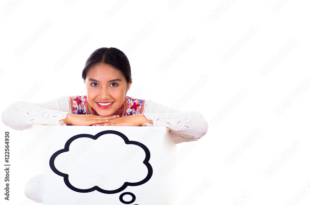 Charming young woman wearing traditional andean blouse with colorful embroideries, matching necklace and earrings, facing camera while posing, leaning over sign which has conversation bubble drawn on Charming young woman wearing traditional andean blouse with colorful embroideries, matching necklace and earrings, facing camera while posing, leaning over sign which has conversation bubble drawn on