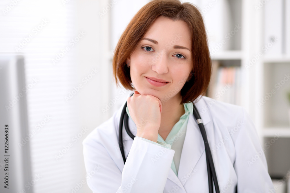 Young brunette female doctor sitting at a desk and working on the computer at the hospital office. Health care, insurance and help concept. Physician ready to examine patient Young brunette female doctor sitting at a desk and working on the computer at the hospital office. Health care, insurance and help concept. Physician ready to examine patient