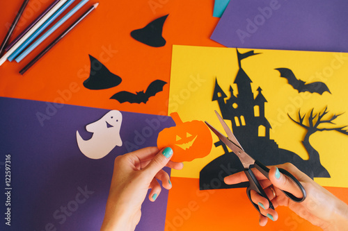 close up of female hands with scissors cut paper orange pumpkin on the background Halloween composition. Castle, ghosts, bats, pumpkins are cut from colored paper. Homemade decorations for Halloween
