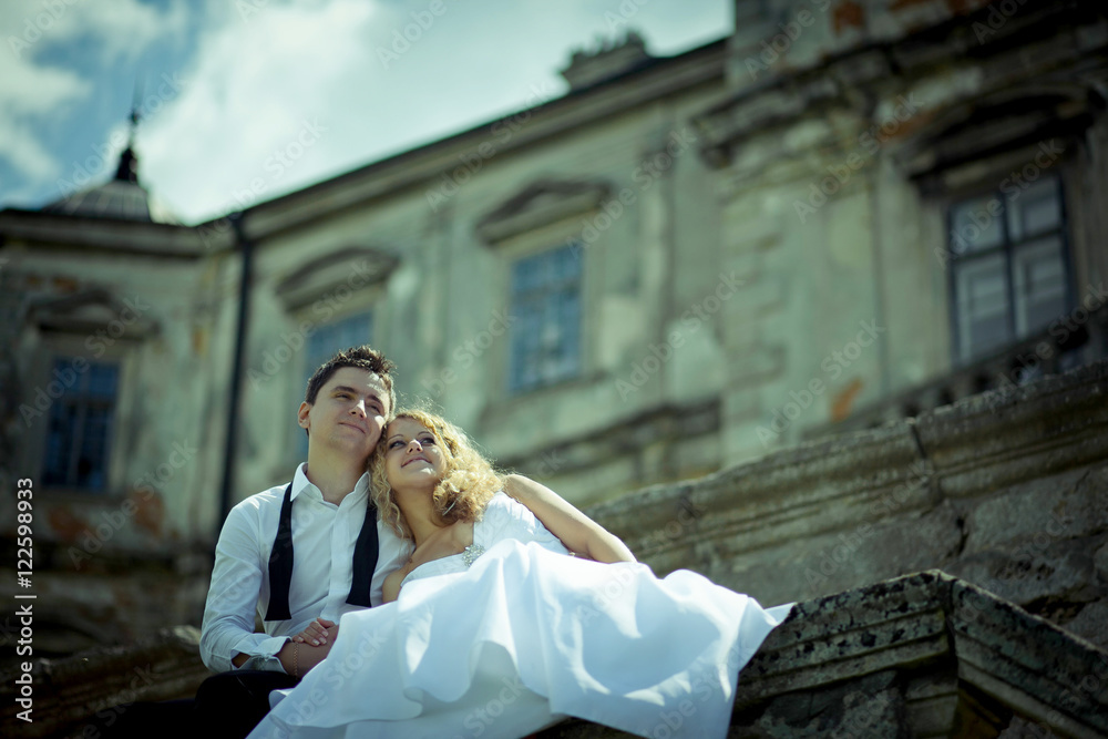 Fototapeta premium Dremy groom hugs his curly bride while they sit on stone handrai