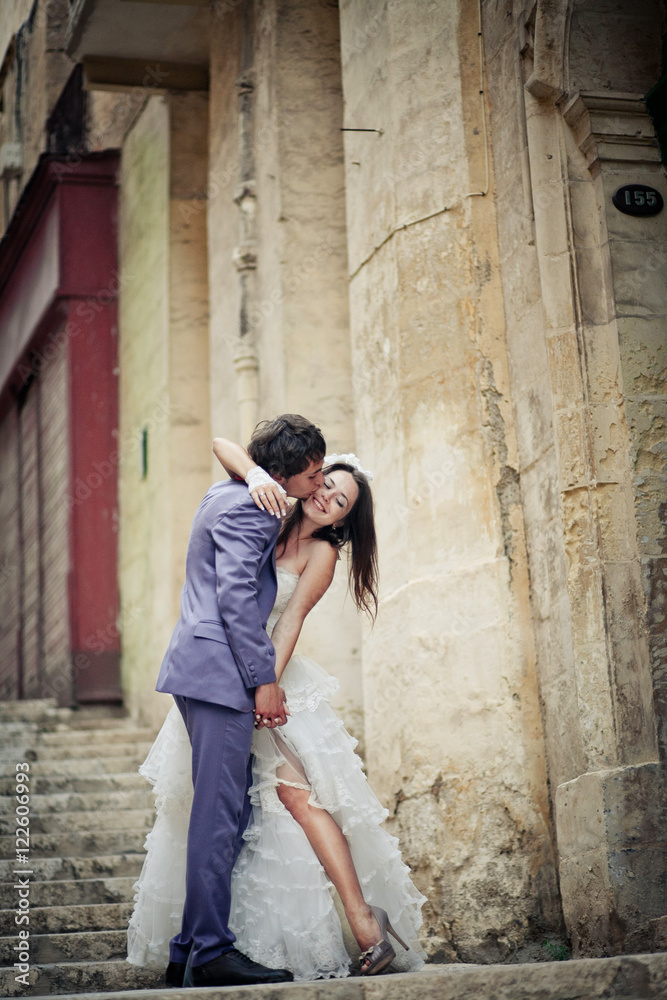 Groom in violet suit kisses bride's cheek while she poses in sed