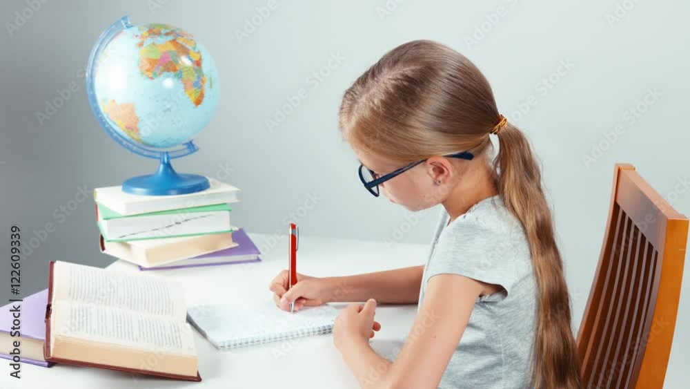 School girl four-eyes writing in her notebook and smiling at camera. Child sitting at desk isolated on white background School girl four-eyes writing in her notebook and smiling at camera. Child sitting at desk isolated on white background