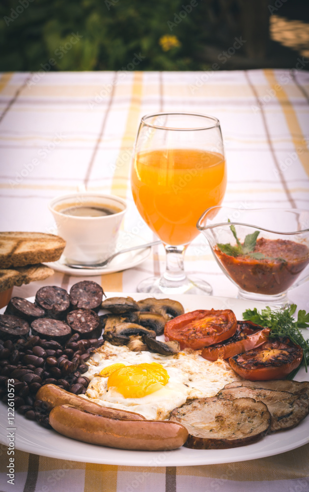 Full English fried breakfast with bacon, egg, sausages, black pudding, mushrooms, grilled tomatoes and baked beans. Closeup with toasts, cup of coffee, orange juice and home made red sauce. Full English fried breakfast with bacon, egg, sausages, black pudding, mushrooms, grilled tomatoes and baked beans. Closeup with toasts, cup of coffee, orange juice and home made red sauce.