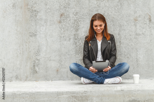 Girl with a tablet and headphones in hands sits on a concrete wall. Online education students © perfectlab