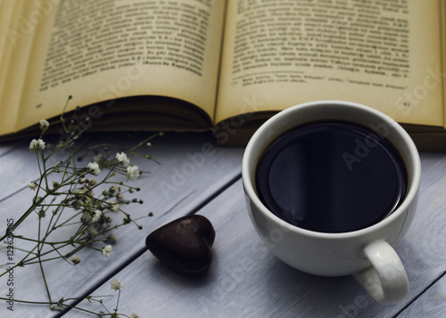 Old book, cup of coffee and heart shaped chocolate on the wooden background