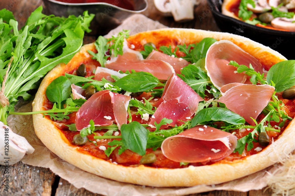 Homemade pizza with tomato sauce, mozzarella cheese, organic arugula, Parma ham, capers and parmesan cheese.