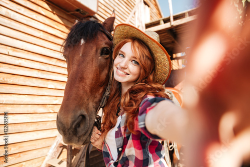 Cheerful woman cowgirl standing taking selfie with horse on farm