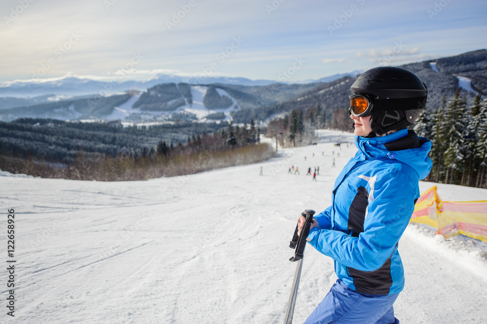 Side portrait of young cute woman skier looking at beautiful mountain landscape at the ski resort on a sunny day. Girl is wearing blue jacket helmet and goggles.