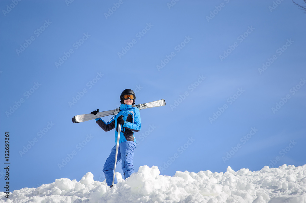 Portrait of active female skier standing on top of the mountain against blue sky on a sunny day. Girl is holding skis on her shoulder smiling and looking into the distance. Winter sports concept. Portrait of active female skier standing on top of the mountain against blue sky on a sunny day. Girl is holding skis on her shoulder smiling and looking into the distance. Winter sports concept.