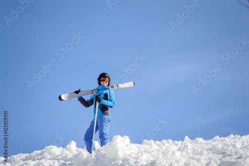 Portrait of active female skier standing on top of the mountain against blue sky on a sunny day. Girl is holding skis on her shoulder smiling and looking into the distance. Winter sports concept.