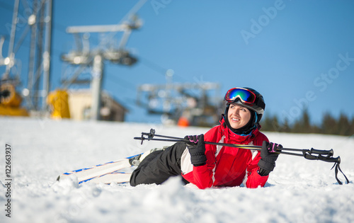 Smiling girl lying with skis on snowy at mountain top, holding ski-sticks and looking away in sunny day at a winter resort with ski lifts and blue sky in background. Close-up