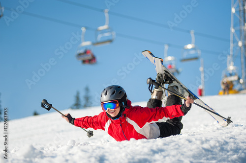 Skier woman in ski goggles lying with raised arms on snowy slope at mountain top in sunny day with ski lifts and blue sky in background. Bukovel, Ukraine