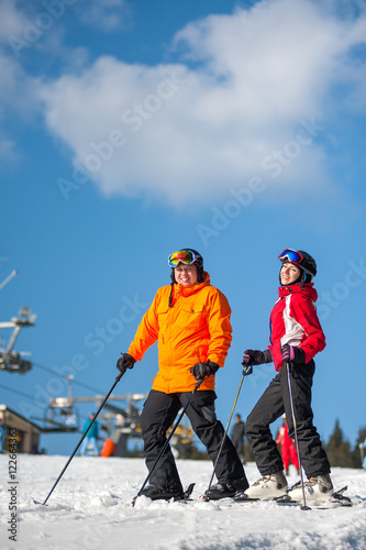 Couple holding skis smiling on mountain top together at a winter resort with ski lifts and blue sky in background. Man is wearing orange jacket, female in red jacket, both is wearing helmets