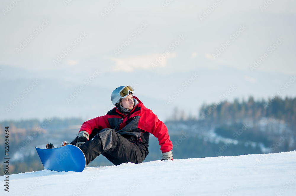 Snowboarder wearing helmet, red jacket, gloves and pants sitting on snowy slope on top of a mountain looking away, with an astonishing view on hills. Carpathian mountains