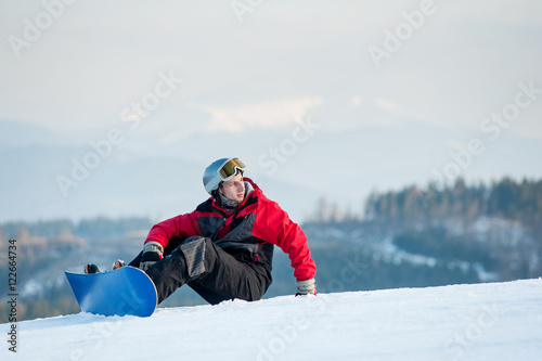Snowboarder wearing helmet, red jacket, gloves and pants sitting on snowy slope on top of a mountain looking away, with an astonishing view on hills. Carpathian mountains