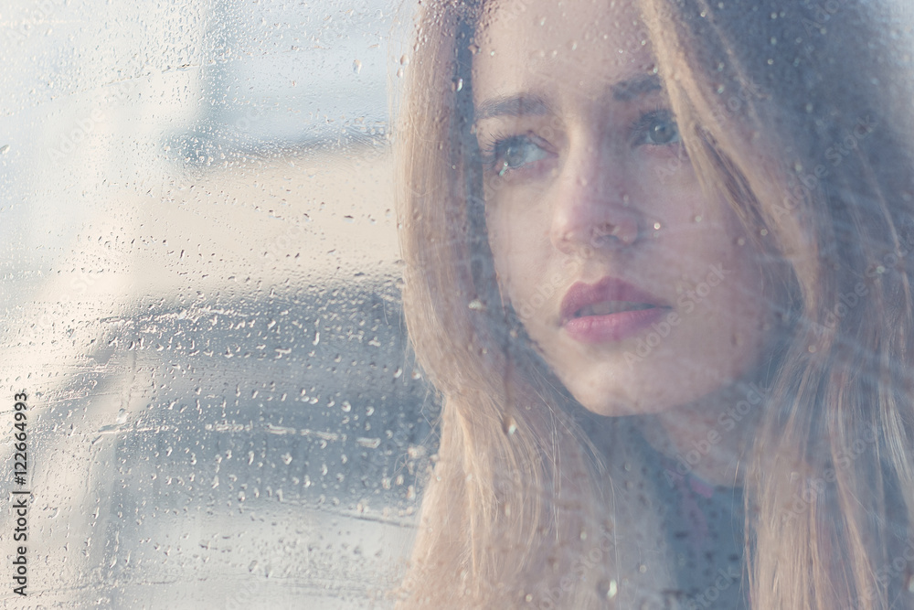 Beautiful sad girl with big eyes in a coat is behind wet glass
