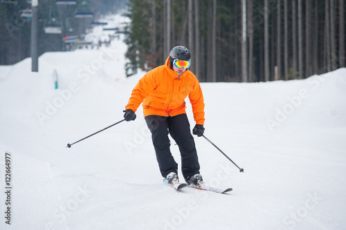 Skier skiing downhill at ski resort against ski-lift and forest. Male is wearing orange jacket, helmet and goggles. Carpathian Mountains, Bukovel