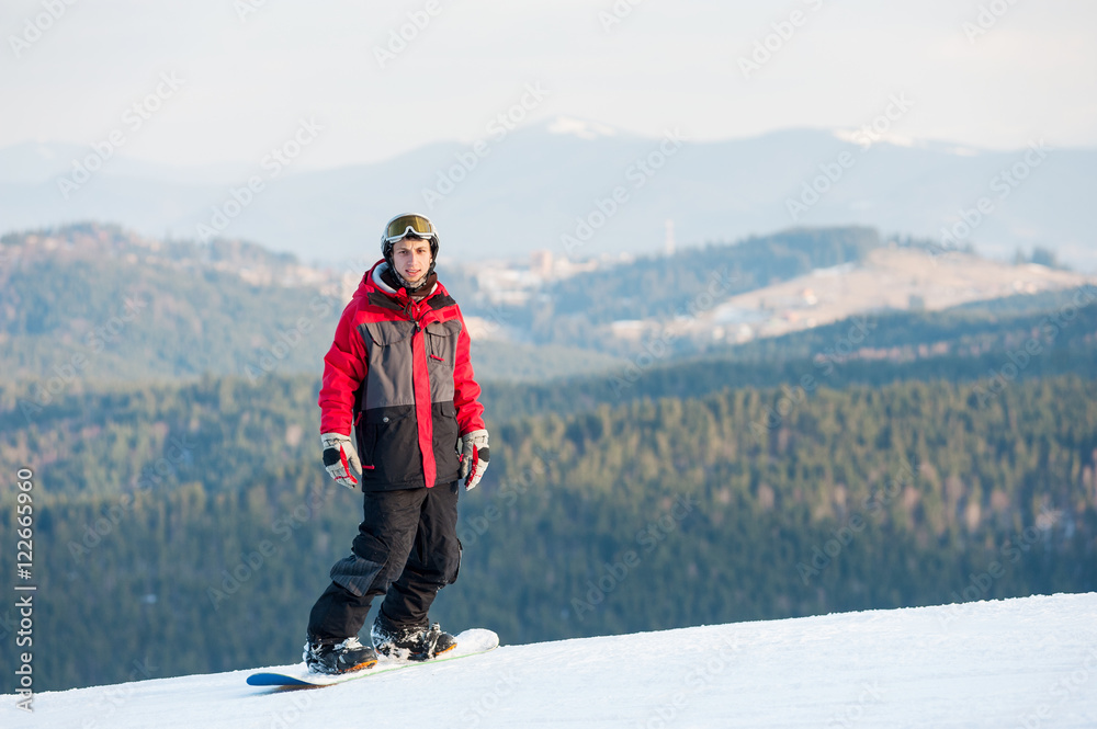 Young snowboarder wearing helmet, red jacket, gloves and pants standing on top of a mountain and looking at the camera on the background of beautiful mountains and forests, winter sports concept