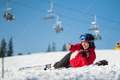 Smiling girl lying with skis on snowy at mountain top, showing thumb up gesture of good class and looking at the camera in sunny day at a winter resort with ski lifts and blue sky in background