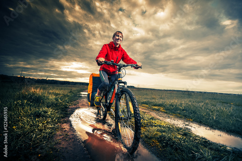 Young lady hiker with loaded bicycle riding through the puddle on a wet rural road