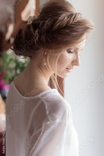 morning beautiful happy young girl of the bride near a window in a white dress with a beautiful boudoir evening festive hairdo with ornaments in their hair and soft makeup