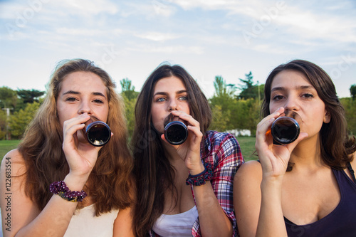 Female friends celebrating the holiday having a good time drinking beer in a park. They re happy. The three girls are drinking at a time.  