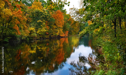 Autumn landscape of a calm river and the wooded shores with colorful foliage and reflection in water on a cloudy day.