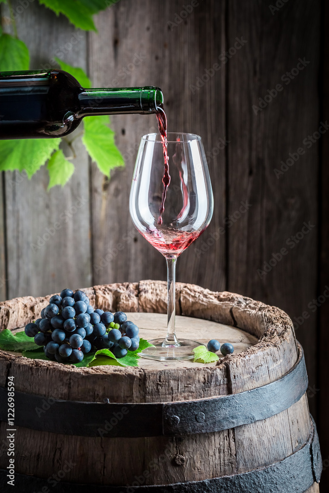Pouring red wine on glass on barrel with grapes