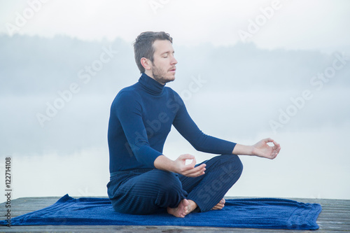 Man relaxing and practicing yoga in the mist on the lake footbridge early morning. Peaceful atmosphere. Foggy air.