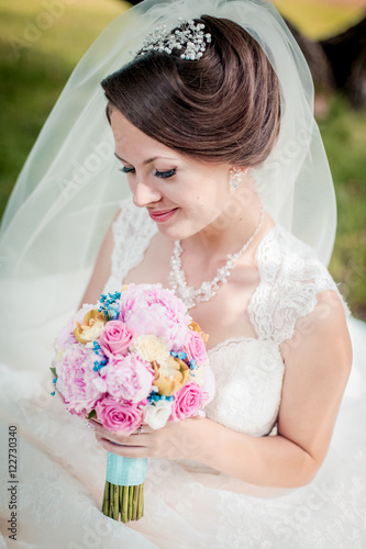 Close up portrait of magical beautiful young bride in elegant white dress with bouquet in the park. Looking at flowers and smiling. Colorful bouquet.