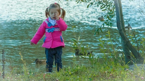 beautiful little girl playing in the park, she is looking at the camera, smiling and waving his hand in greeting . ducks on the lake.