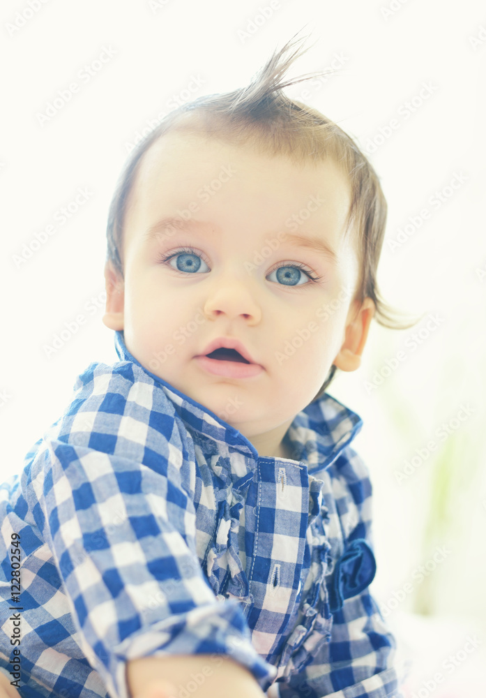 A cute little baby is sitting and looking into the camera and is wearing a blue shirt. The baby could be a boy or girl and has blue eyes. Use it for a parenting or love concept. A cute little baby is sitting and looking into the camera and is wearing a blue shirt. The baby could be a boy or girl and has blue eyes. Use it for a parenting or love concept.