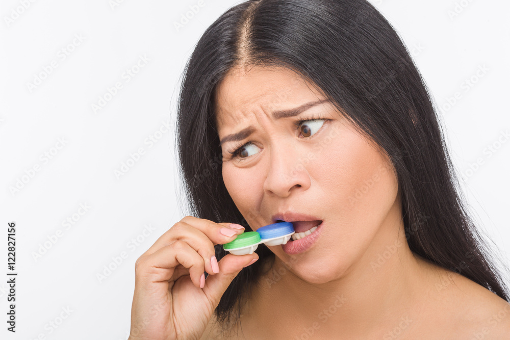 Korean or Asian woman biting container for contact lenses isolated on white background. Attractive lady with brunette hair in studio. Studio shot. Korean or Asian woman biting container for contact lenses isolated on white background. Attractive lady with brunette hair in studio. Studio shot.