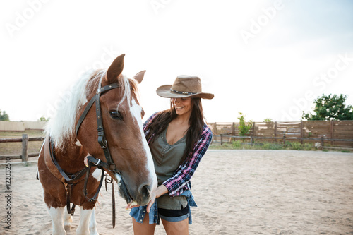 Cheerful woman cowgirl standing and taking care of her horse © Drobot Dean