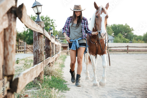 Woman cowgirl in hat walking with her horse in village photo