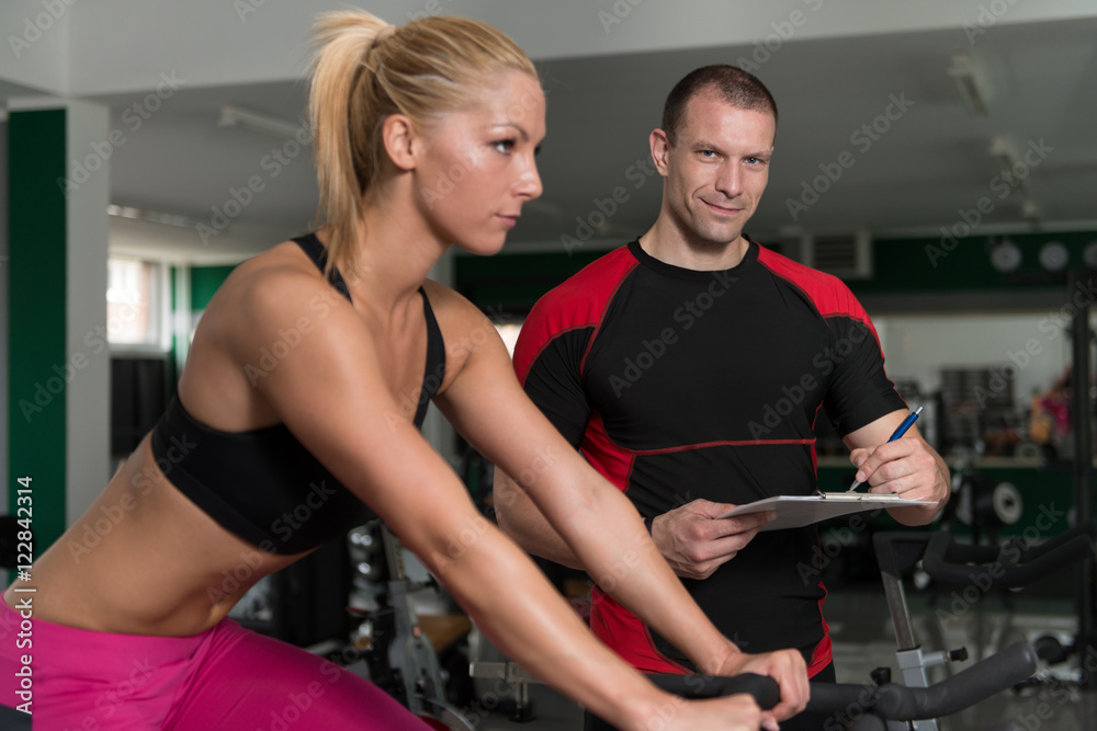 Woman Train Bicycle On Machine With Personal Trainer