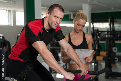 Man Train Bicycle On Machine With Personal Trainer