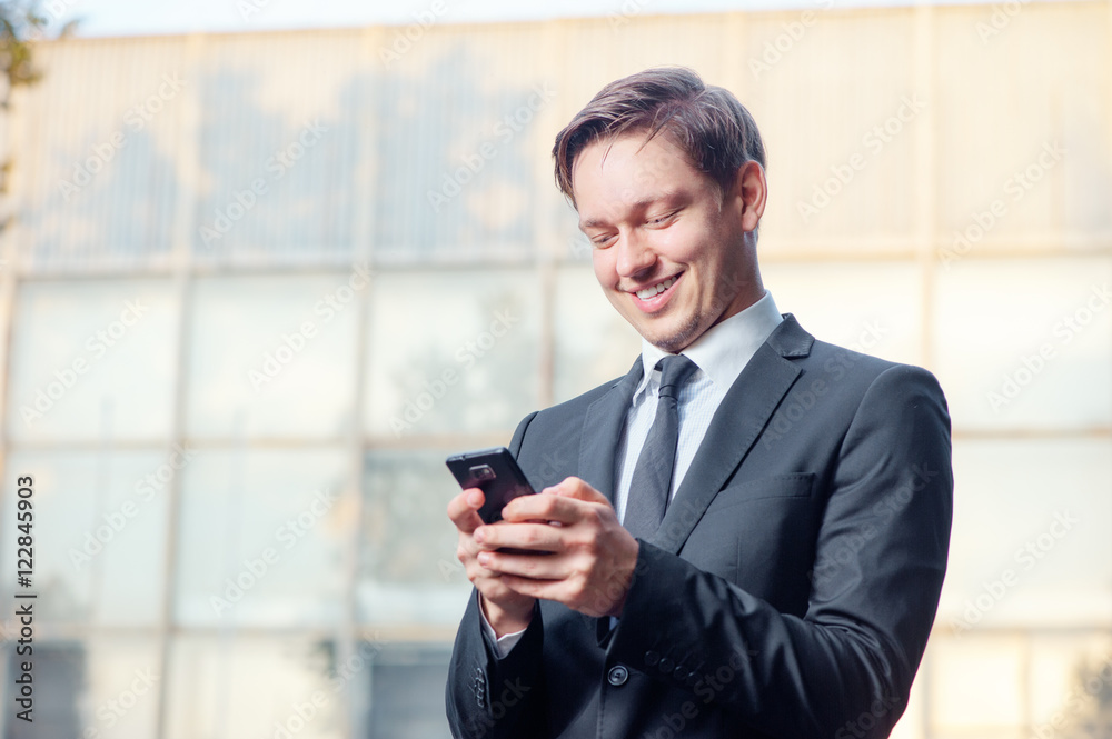 Typing business message. Cheerful young man in formal wear holding mobile phone and smiling while standing outdoors Typing business message. Cheerful young man in formal wear holding mobile phone and smiling while standing outdoors