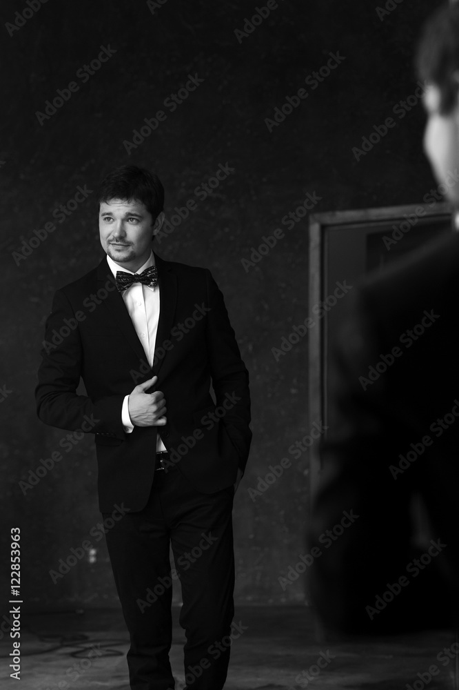Young handsome stylish man in white jacket and black formal suit with bow-tie standing in interior and posing against mirror Young handsome stylish man in white jacket and black formal suit with bow-tie standing in interior and posing against mirror