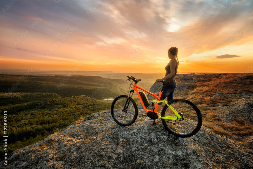 Sunset from the top /
A woman with a bike enjoys the view of sunset over an autumn forest