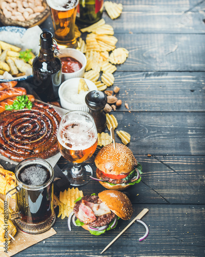 Beer and snack set. Octoberfest food frame concept. Variety of beers, grilled sausages, burgers, fried potato, corn, chips and sauces on dark wooden scorched background. Selective focus, copy space