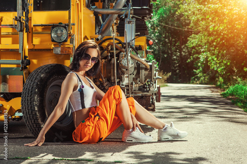 Sexy woman worker in overalls is sitting near the tractor.