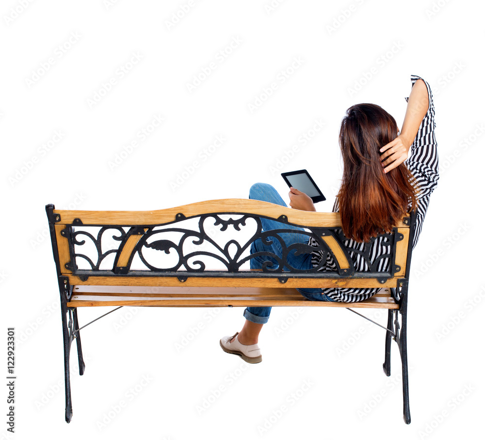 back view of woman sitting on bench and looks at the screen of the tablet.  Rear
