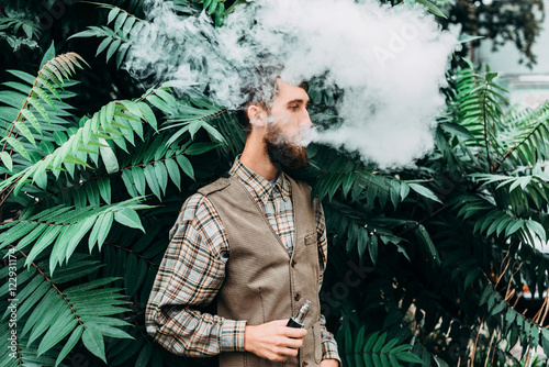 Tableau sur toile Young man with beard vaping an electronic cigarette outdoor