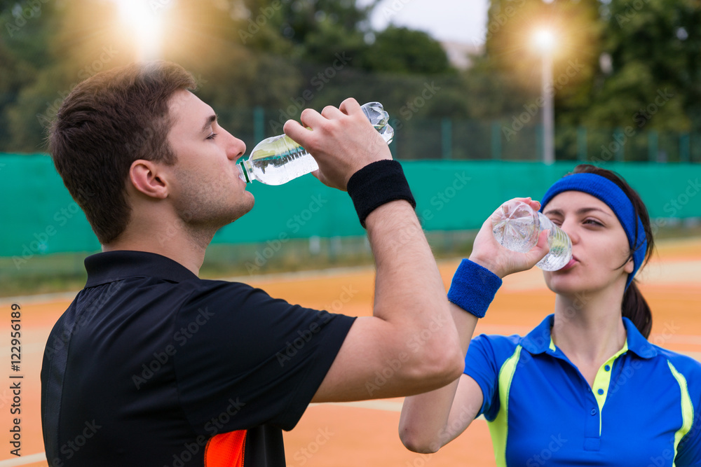 Young couple of tennis players drinking water on clay court outd