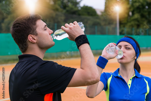 Young couple of tennis players drinking water on clay court outd